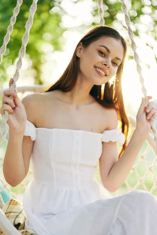 Young woman with long hair wearing a white off shoulder dress, sitting on a swing in a sunny garden, exuding joy and relaxation