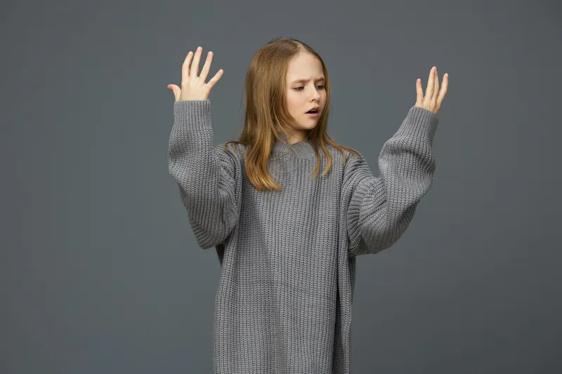 Young woman in oversized gray sweater expressing frustration with raised hands against a minimalist gray background, showcasing deep emotions and contemporary fashion