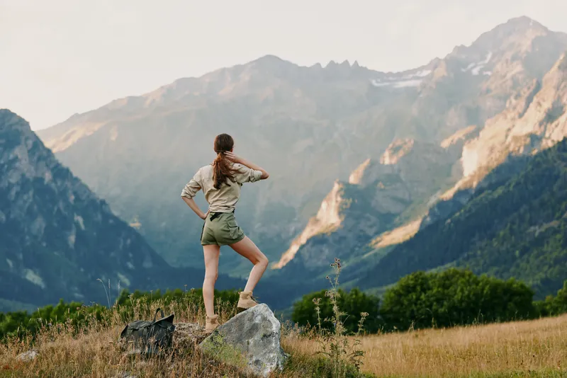 A woman in casual clothes stands on a rock looking at mountain peaks during daytime. The scene shows outdoor adventure, nature exploration, and lifestyle in a natural setting.