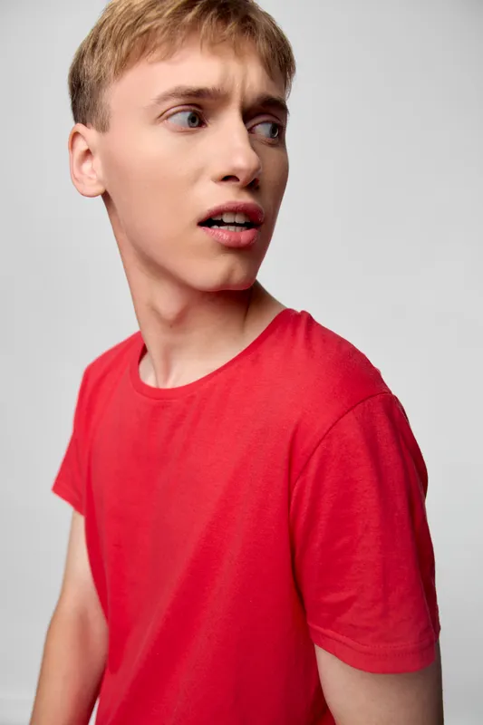 Boy with surprised expression wearing a red shirt in a studio setting, looking to the side, casual pose for lifestyle and fashion stock use.
