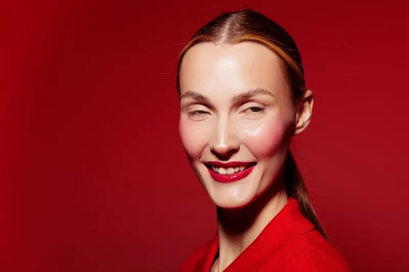 Smiling woman with slicked hair and red lipstick wears red jacket against red background. Studio portrait with bright makeup and confident expression. Concept of fashion, beauty, and style.