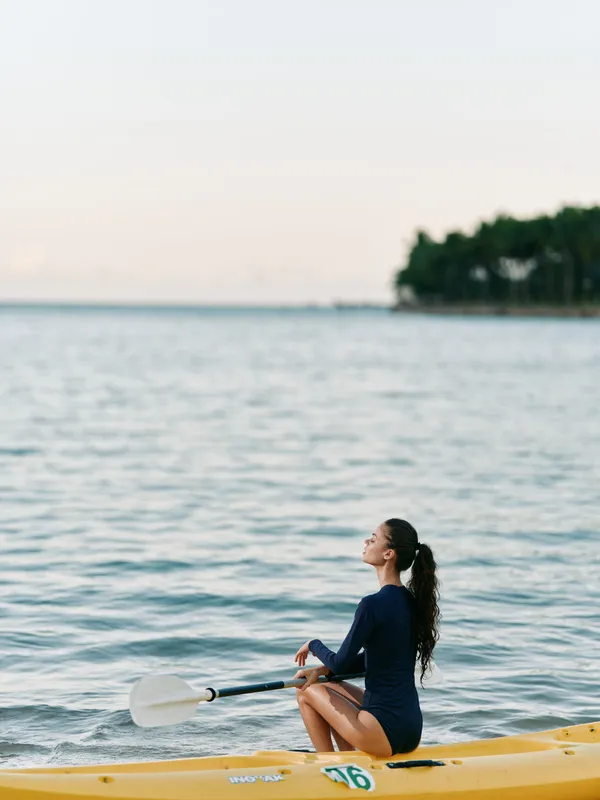 A lone kayaker sits calmly on a calm sea in a dark swimsuit, paddle resting beside the kayak, distant trees on the horizon, and tranquil light creating a peaceful coastal mood.