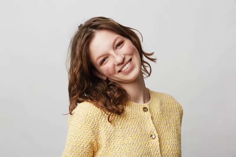 Smiling young woman with curly brown hair wearing a yellow knitted sweater poses against a light grey background. Concept of happiness, casual style, and natural beauty.