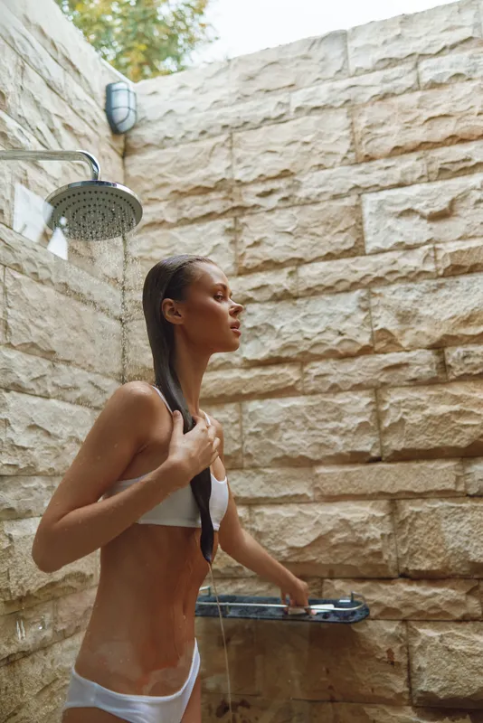 woman showering outdoors with stone wall background, wet hair, natural light, healthy skin, summer wellness and self-care routine