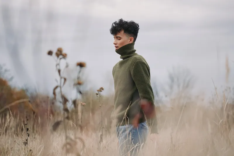 A contemplative young man in a green turtleneck stands in a windswept field, captured in an authentic, natural moment that emphasizes genuine presence and honest portrait storytelling.