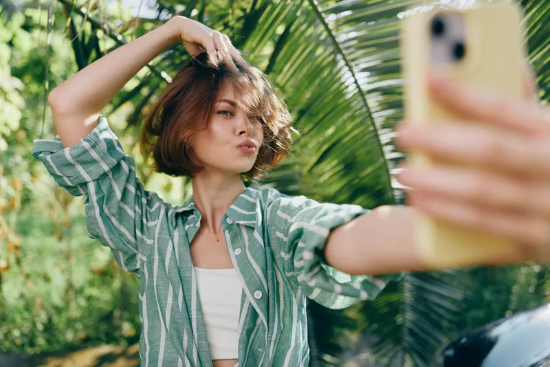 woman selfie outdoors smiling with casual attire; a cheerful portrait captured under natural light; she poses gracefully, creating a vibrant, relaxed mood amid lush greenery and bright sunshine