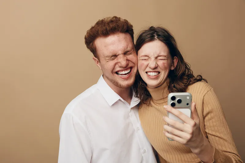 Happy young couple with closed eyes laughing together while taking a selfie on smartphone. Casual clothing and warm beige background create a cozy and joyful atmosphere.