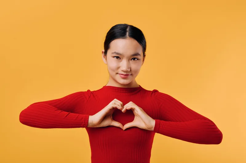 Young woman wearing red sweater making heart shape with hands in front of orange background for love, care, support, friendship, and positive emotions concept.