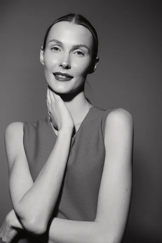 Portrait of a slender Caucasian woman with fair skin and sleek hair, wearing a sleeveless top, posing against a plain background in black and white.