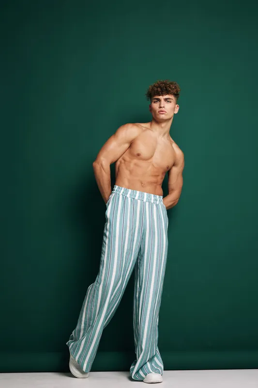 shirtless handsome man with curly hair in striped loose pants and sneakers posing against green neutral background light for style and fashion photography portrait