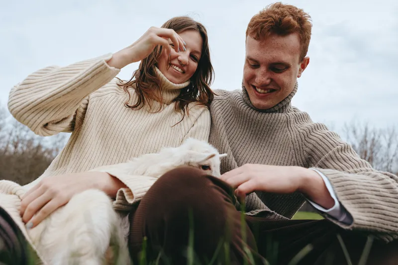 Casual couple outdoors with a curious goat kid, sharing a light moment in a sunlit field. A lifestyle scene that captures warmth, affection, and playful interaction with animals in a natural setting.