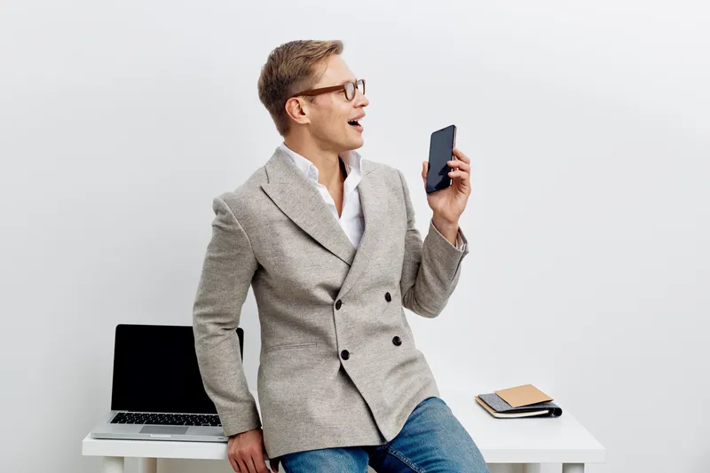 young man with smartphone talking online smile casual wear jacket glasses sit on desk laptop notebook white background remote work digital communication concept