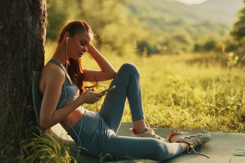 Young athletic woman relaxing outdoors in activewear, enjoying music, green nature background, exploring healthy lifestyle and fitness concepts