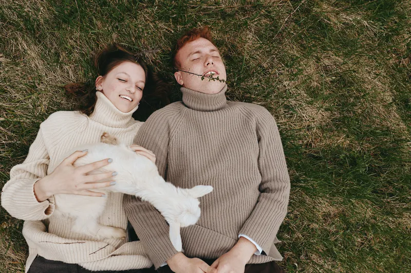 Joyful couple lying on grass in cozy sweaters, cuddling a small white dog, relaxed lifestyle moment outdoors, warm tones, natural light, connection and comfort in a casual romantic scene