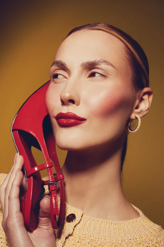 Close-up of a stylish woman with red lipstick holding a red high heel shoe near her face. Concept of fashion, beauty, glamour, and elegance on yellow background.