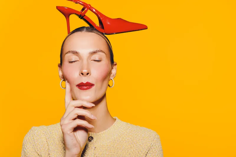 Young woman with closed eyes and red lipstick holds red high heel shoe on head against bright yellow background. Concept of fashion, style, beauty, and creativity.