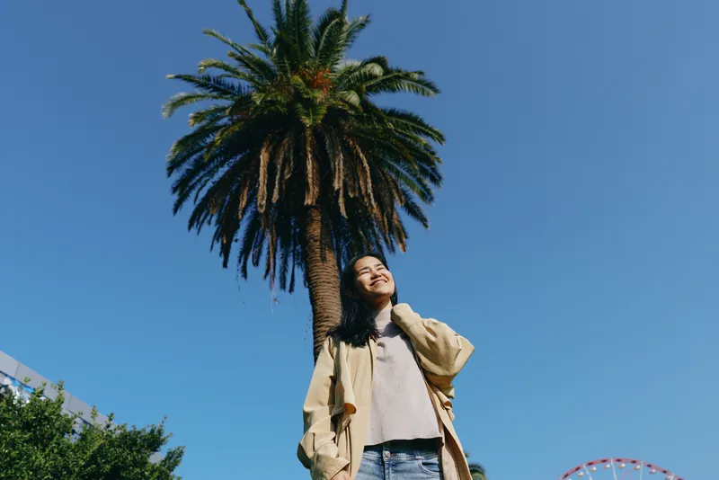Palm tree against a bright sky, woman looking up, wearing a beige coat and denim, street fashion on a sunny day, urban mood and confident style in motion, playful and cinematic vibe