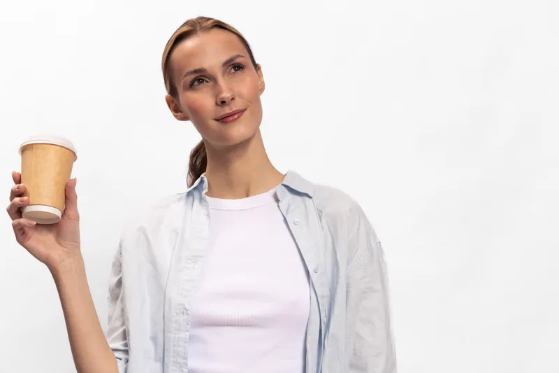 Young woman with ponytail holding disposable coffee cup looks away with slight smile. Casual outfit and neutral background create relaxed and thoughtful mood. Concept of coffee break and leisure.