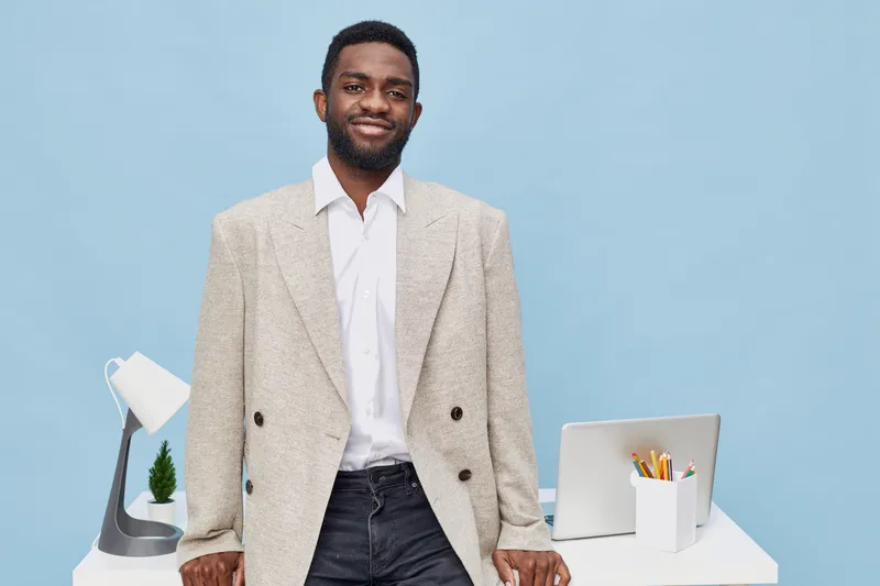 Smiling man standing behind a desk with a laptop, desk lamp, and stationery in a modern office setting, wearing a beige blazer and white shirt, friendly professional appearance, workspace environment.