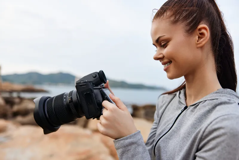 Young female photographer in a casual gray hoodie smiles while adjusting her camera on a rocky beach, reflecting creativity and passion for photography