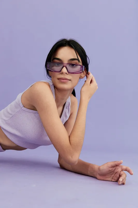 young woman wearing purple sunglasses and purple top poses on pastel purple background with relaxed expression, sleek black hair, and minimal makeup in high-key lighting