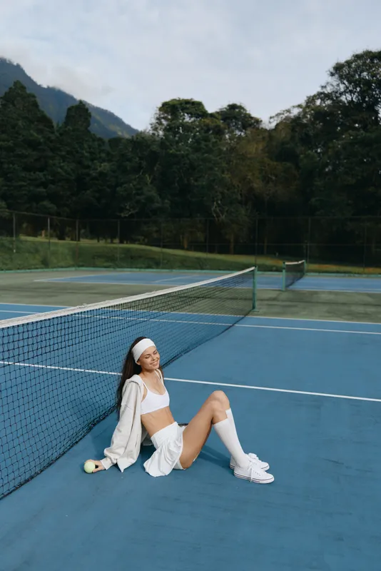 Young female athlete relaxing on a tennis court, dressed in sporty white outfit, smiling with confidence, surrounded by green nature and blue court