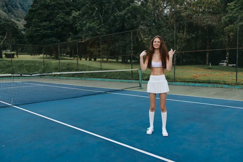 Young female tennis player in sporty attire shows positivity on blue court with greenery, wearing a white crop top and skirt, perfect for summer sports activity