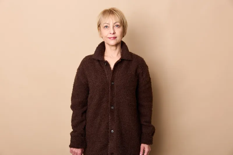 Elderly woman portrait, beige studio background, dark coat, standing figure, winter comfort, calm expression, soft studio lighting, concept of aging, wellness, dignity