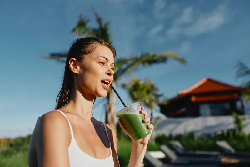 Young woman in a white tank top drinks a vibrant green smoothie by a sunny poolside, enjoying a refreshing moment with palm trees and a clear blue sky behind, a gentle breeze ruffles her hair
