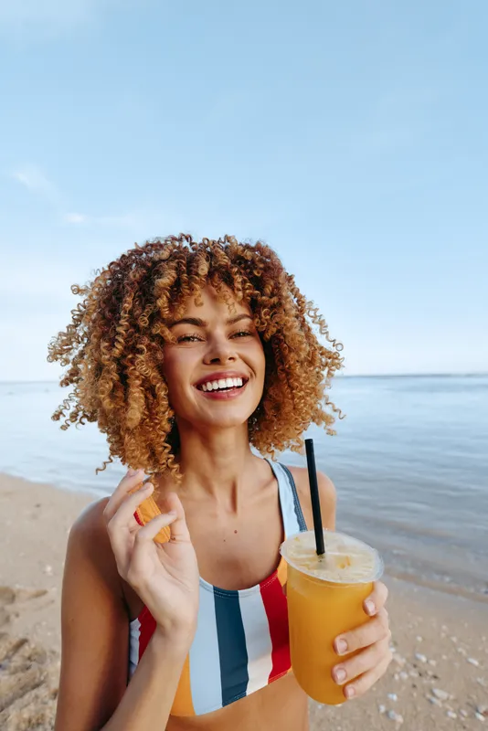 Young woman with curly hair laughs on a sunny beach, holding a bright orange juice and wearing a striped swimsuit, with a calm sea behind and a light breeze.
