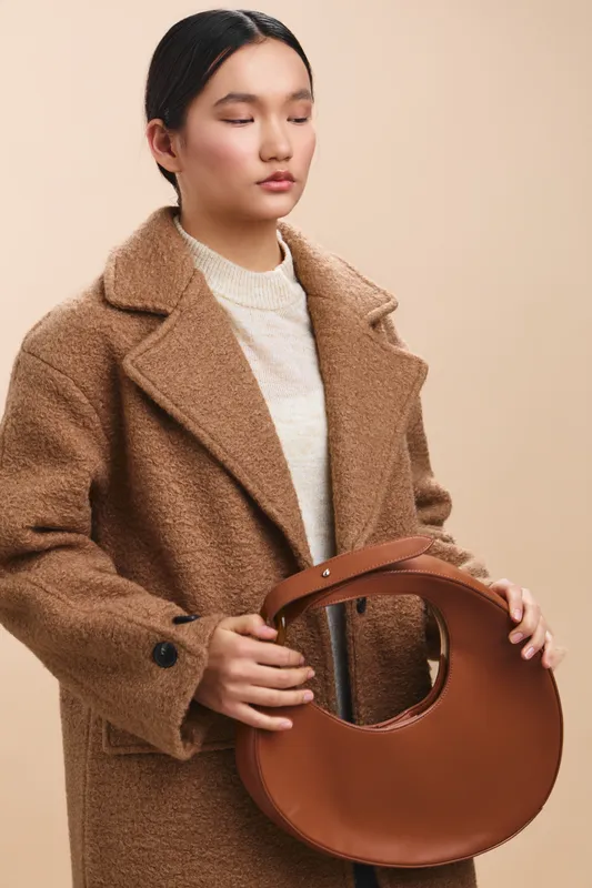 Asian woman stands in a neutral studio wearing a warm brown coat and holding a circular leather bag, presenting a calm, minimalist fashion moment.