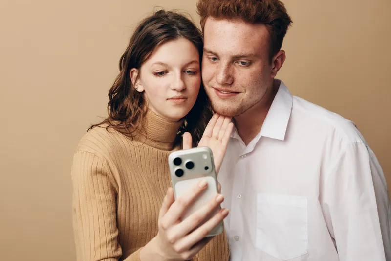 Young couple with casual clothes looking at smartphone screen together, sharing moment of connection and technology use in neutral studio background. Concept of communication and relationship.