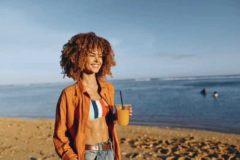 A smiling woman on a sunny beach holds a yellow drink, wearing an orange shirt and striped bikini top. A casual summer moment captured in natural light, highlighting coastal style.