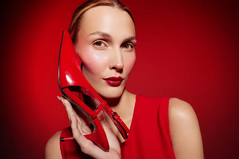Close-up of a woman with red lipstick holding a red high heel shoe near her face. The red background and dress create a bold fashion concept with strong color contrast and style.