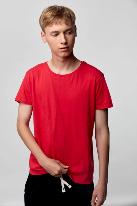 Red tee, young man in casual fashion poses in studio with a relaxed stance, hands near drawstring, plain background, modern wardrobe suitable for everyday wear and street style.