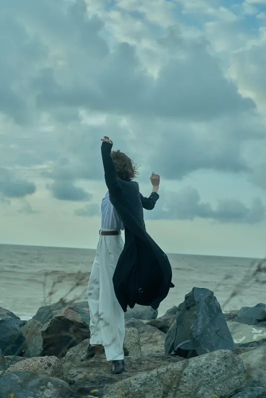 Windy shoreline scene with a figure in layered clothing raising arms on a rocky jetty, embracing the open sea and distant horizon, under a dramatic sky.