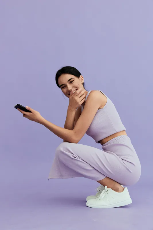 Smiling woman in casual outfit squatting on violet background, holding remote control, wearing white sneakers, showing playful and relaxed mood in studio setting.