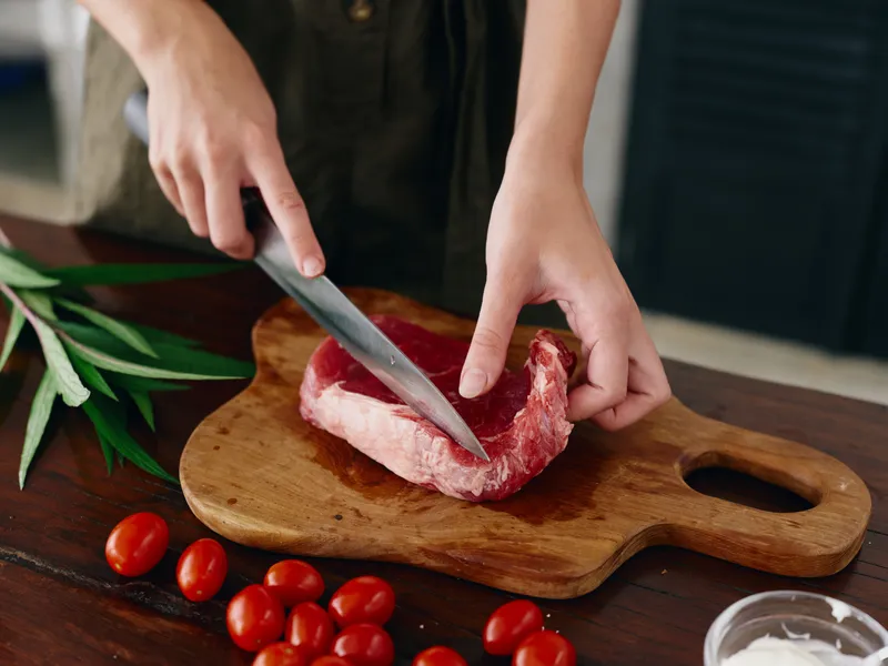 Женщина с ножом в руке нарезает свежее мясо стейка для жарки на кухне, на столе соль, перец и другие специи, красные помидоры черри и зелень, готовит ужин