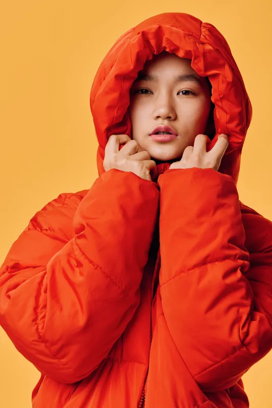 Woman wearing orange hooded jacket with hands pulling hood close, portrait in studio with yellow background. Female model with natural makeup and calm expression in warm clothing for cold weather.