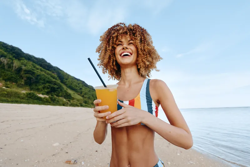 Woman with curly hair smiling on beach holding a cold drink with a straw, summer vacation seaside mood, outdoor sunshine, relaxed healthy refreshment near ocean waves and green hills