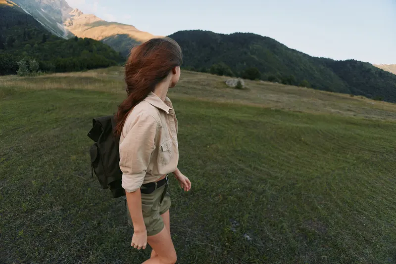 Woman hiker stands in an open field with a backpack, gazing toward distant mountains, capturing a lifestyle moment of outdoor adventure in a vast natural landscape