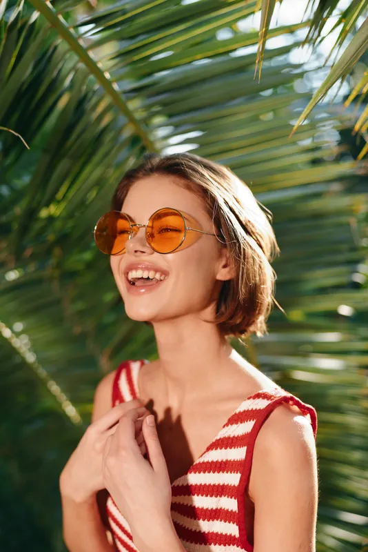 A cheerful woman wearing a striped sleeveless top and orange round sunglasses smiles brightly under sunny palm fronds, enjoying a carefree tropical moment.