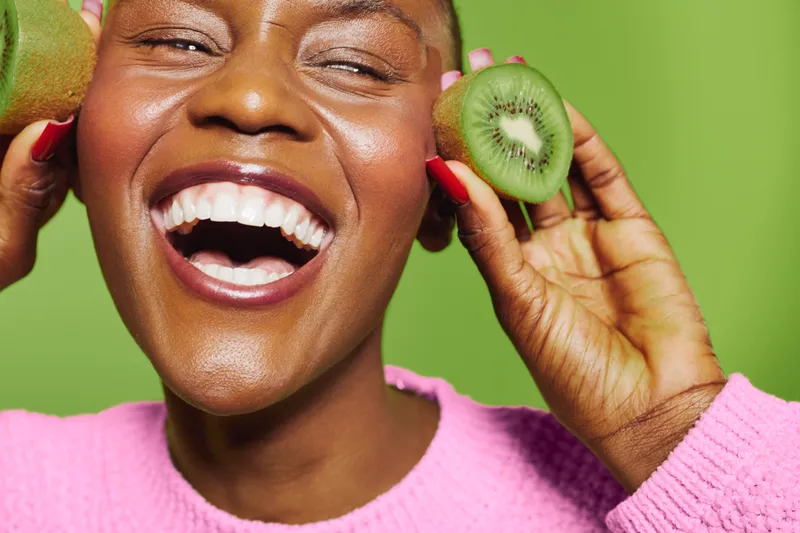 Happy African woman with dark skin laughing and holding kiwi fruit slices near her face. Close-up on green background. Concept of healthy lifestyle, fruit, and natural beauty.