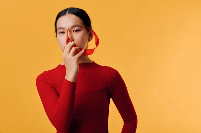 Woman in red sweater posing with heart-shaped object against orange background. Studio portrait of female model with calm expression and long dark hair tied back in minimalist style.