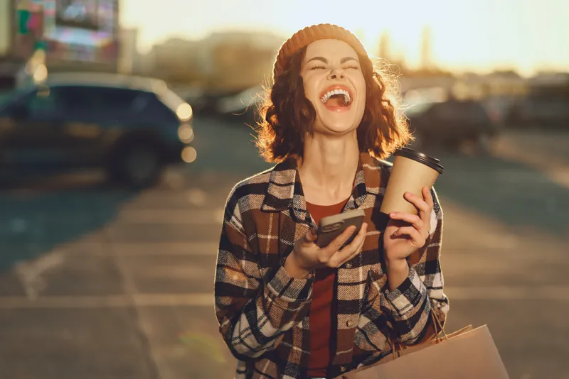 Woman laughter coffee phone shopping beanie sunlight street captured in candid lifestyle moment; authenticity and emotional storytelling during golden hour glow for mindful living urban joy.