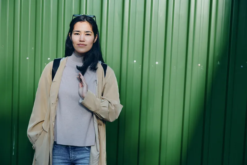 Woman in beige coat and gray scarf stands against a green metal wall, casual street style with urban flair, ready for modern fashion and lifestyle photography projects