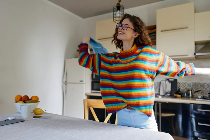 Smiling woman wearing a colorful striped sweater dancing beside a kitchen table, enjoying a cozy home moment. Bright daylight, warm mood, everyday living and wellbeing.