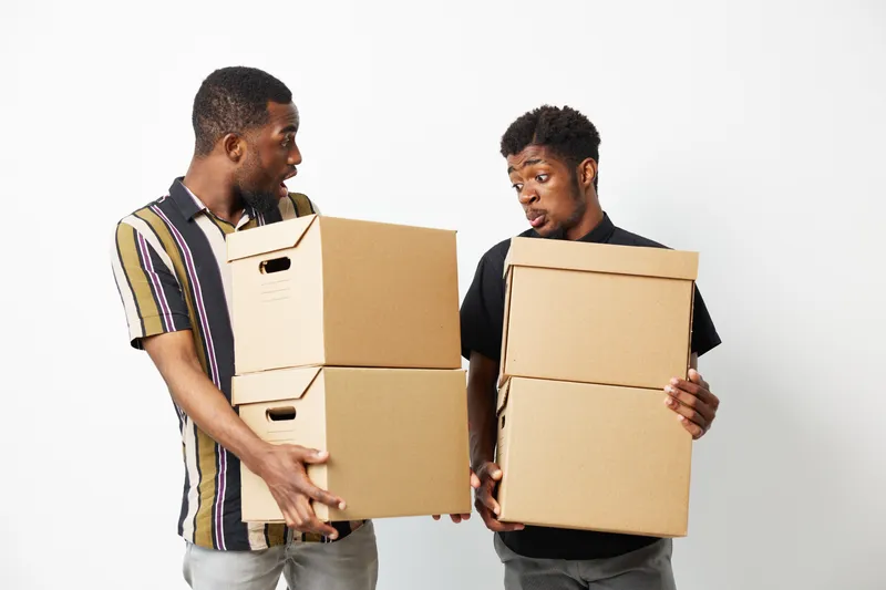 Two black men sharing a surprised moment while carrying cardboard boxes, showcasing their teamwork and excitement for a new project together.