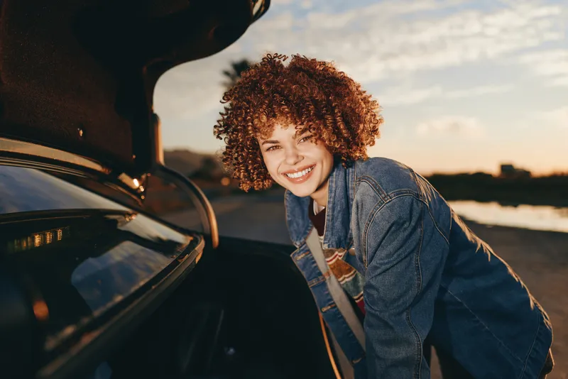 woman lifestyle outdoors at sunset, curly hair, denim jacket, smiling beside car, relaxed travel moment, natural sunlight, roadside scenery, with expression and open road feel