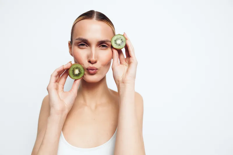 Young woman holding two halves of fresh kiwi fruit near her face, playful expression, clean white background, natural light, healthy lifestyle concept, minimalistic style